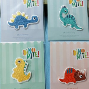 May include: Four colourful iron-on patches featuring different dinosaurs. Each patch has a different dinosaur with a cute expression and a short message about making the world a better place. The patches are labelled with the dinosaur's name: Stegosaurus, Brontosaurus, Tyrannosaurus Rex, and Triceratops.