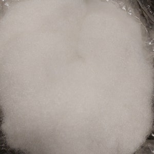 Op de afbeelding: Een close-up van een grote hoeveelheid witte polyester vulling, die vaak wordt gebruikt voor het vullen van kussens, kussens en speelgoed. De vulling zit in een doorzichtige plastic zak.