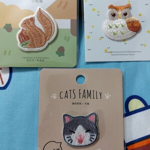 Può includere: Tre toppe da stirare raffiguranti uno scoiattolo, un gufo e un gatto. La toppa dello scoiattolo è marrone e presenta il testo "森之暖暖 小松鼠" in nero. La toppa del gufo è bianca con accenti marroni e arancioni e presenta il testo "Decoration Sticker" in nero. La toppa del gatto è grigia e bianca con il testo "CATS FAMILY 喵呜家族/灰囉" in nero. Tutte e tre le toppe presentano il testo "Cardlover & Embroidery 信的恋人刺绣贴" in nero.