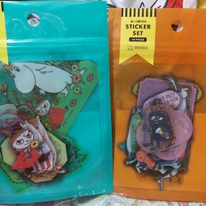 May include: Two clear plastic bags with colorful sticker sets inside. The sticker sets feature cartoon characters and illustrations. One bag has a sticker set with a Moomin character and the text "You felt needed for me". The other bag has a sticker set with a character from the cartoon "Moomin" and the text "I won't felt needed for me".