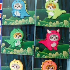 Könnte beinhalten: Sechs gestickte Bügelbilder mit einer Katze, die ein Dinosaurierkostüm trägt. Die Patches befinden sich auf einem grünen Hintergrund mit einer Waldszene. Die Dinosaurier sind blau, grün, pink, gelb und orange.