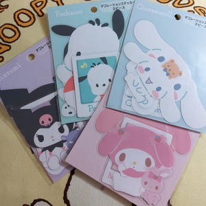 Könnte beinhalten: Ein Set aus vier dekorativen Aufklebern mit beliebten japanischen Comicfiguren. Die Blätter sind rosa, lila, blau und weiß. Die Figuren sind Kuromi, Pochacco, Cinnamoroll und My Melody. Jedes Blatt trägt den Text "デコレーションステッカー", was "Dekorationsaufkleber" bedeutet.