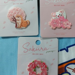 May include: Three pink and white cards with embroidered patches. The patches feature a cherry blossom tree with a dog, a cat sitting on a cloud of cherry blossoms, and a wreath of cherry blossoms. The cards have the text "Sakura series" and "Cardlover & Embroidery 信的恋人刺绣贴" in Japanese and Chinese characters.