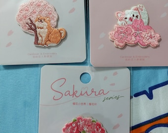 May include: Three pink and white cards with embroidered patches. The patches feature a cherry blossom tree with a dog, a cat sitting on a cloud of cherry blossoms, and a wreath of cherry blossoms. The cards have the text "Sakura series" and "Cardlover & Embroidery 信的恋人刺绣贴" in Japanese and Chinese characters.