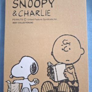 Peut inclure: Bloc-notes à spirales avec Snoopy et Charlie Brown de la bande dessinée Peanuts. La couverture est beige avec des dessins au trait noirs des personnages et le texte "SNOOPY & CHARLIE". Le bloc-notes est de Best Collection, Inc.