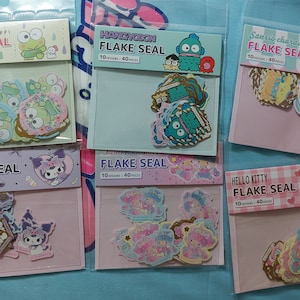 Op de afbeelding: Zes pakjes met kleurrijke karakter stickers. Elk pakje heeft een ander karakter, waaronder Keroppi, Hangyodon, Sanrio-karakters, Kuromi, Little Twin Stars en Hello Kitty. Elk pakje heeft een roze of blauwe achtergrond met de naam van het karakter en de tekst "FLAKE SEAL" erop gedrukt.