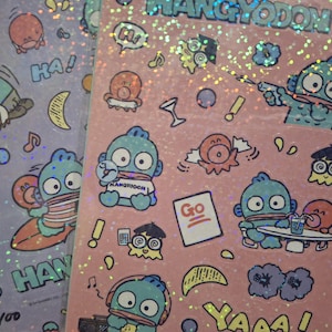 Op de afbeelding: Een vel met kleurrijke stickers met een cartoonfiguur genaamd Hangyodon. De stickers zijn op een roze achtergrond met glitter. De stickers tonen het personage in verschillende poses en met verschillende uitdrukkingen. De stickers bevatten ook tekst zoals "Hoi!" en "Ga!"