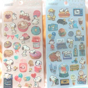 Könnte beinhalten: Zwei Bögen Aufkleber zum Thema Peanuts. Ein Bogen zeigt Snoopy mit Lebensmitteln wie Donuts und Kaffee. Der andere Bogen zeigt Snoopy mit einem Fernseher, einer Schreibmaschine und anderen Gegenständen. Die Aufkleber sind farbenfroh und haben ein glitzerndes Finish.