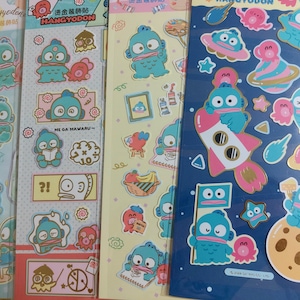 Op de afbeelding: Drie vellen met stickers met een cartoonfiguur genaamd Hangyodon. De stickers zijn kleurrijk en hebben verschillende ontwerpen, waaronder een raket, een planeet en een vis. De tekst op de stickers luidt "Hangyodon" en "Me Ga Mawaru".