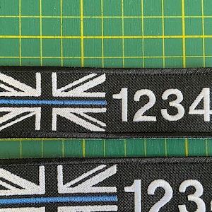 May include: Two black fabric patches with a white embroidered Union Jack flag and a thin blue line across the flag. The number 1234 is embroidered in white below the flag on each patch.