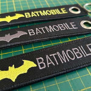 May include: Three black fabric keychains with a silver metal ring. Each keychain has a bat silhouette and the word "BATMOBILE" embroidered in yellow, gray, or silver thread.