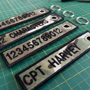 May include: Four keychains with black text on a camouflage background. The keychains feature the numbers "123456789012", "12 CHARACTERS", "123456789012", and "CPT HARVEY".
