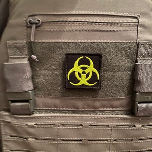 Könnte beinhalten: Eine graue taktische Weste mit einer schwarzen Reißverschlusstasche und einem gelben Biohazard-Symbol-Patch.