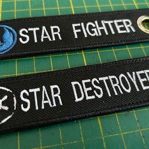 May include: Two black fabric keychains with white embroidery. The top keychain says "STAR FIGHTER" with a blue Rebel Alliance symbol. The bottom keychain says "STAR DESTROYER" with a white Imperial symbol.
