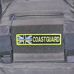 May include: A grey tactical vest with a black zipped pocket and a black and white patch with the text "COASTGUARD" in yellow.