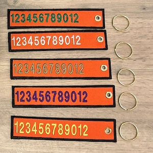 May include: Five orange keyrings with black trim and a metal ring. Each keyring has the numbers 123456789012 embroidered in different coloured thread: green, white, gold, purple, and yellow.