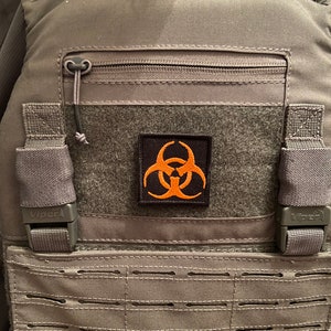 May include: A grey tactical vest with a black zipper and a black and orange biohazard symbol patch. The vest has adjustable straps and a MOLLE system.