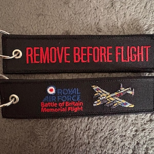 May include: Two rectangular black fabric keychains with silver metal rings. The top keychain features the red embroidered text REMOVE BEFORE FLIGHT. The bottom keychain displays the Royal Air Force Battle of Britain Memorial Flight logo and a plane.