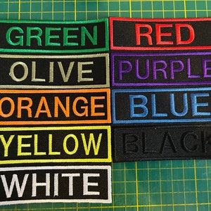 以下が含まれることがあります： 緑、赤、紫、青など、さまざまな色の刺繍パッチのコレクション。各パッチは、黒い背景に大文字で色名を表示しています。パッチは、緑色のカッティングマットに配置されています。