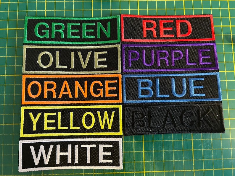 以下が含まれることがあります： 緑、赤、紫、青など、さまざまな色の刺繍パッチのコレクション。各パッチは、黒い背景に大文字で色名を表示しています。パッチは、緑色のカッティングマットに配置されています。