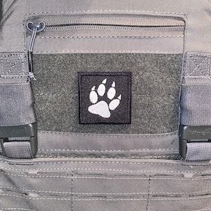 May include: A grey tactical vest with a black patch featuring a white paw print. The vest has adjustable straps and a zipped pocket.