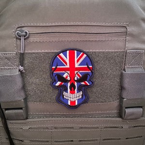 Pode incluir: Um colete tático cinza com um remendo de caveira com a bandeira da Union Jack. O remendo é costurado na frente do colete.