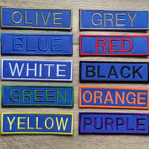 Puede incluir: Doce parches rectangulares de tela de diferentes colores con los nombres de los colores bordados en hilo blanco. Los colores son oliva, gris, azul, rojo, blanco, negro, verde, naranja, amarillo y morado.