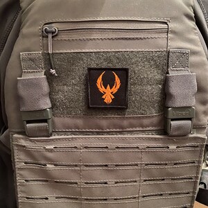 May include: A gray tactical vest with a black zippered pocket and a black patch with an orange logo. The vest has a black MOLLE panel with multiple rows of webbing.