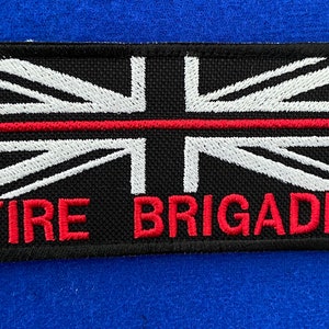 May include: A black and white embroidered patch with a red stripe across the middle. The patch features a Union Jack flag design and the words "FIRE BRIGADE" in red.