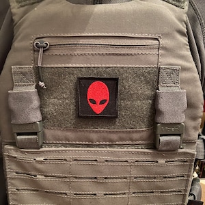 Pode incluir: Um colete tático cinza com um patch de alienígena vermelho na frente. O colete tem um bolso com zíper e alças ajustáveis.
