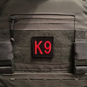 Op de afbeelding: Een zwarte tactische vest met een rode K9-patch op de voorkant. De vest heeft een ritszak en verstelbare banden.