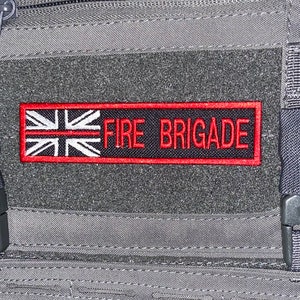 Pode incluir: Um colete tático cinza com um remendo preto que diz "FIRE BRIGADE" com uma bandeira da União Jack vermelha e branca no lado esquerdo.