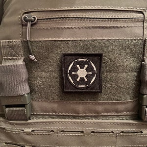 May include: A gray tactical vest with a black zippered pocket and a black patch with a white symbol. The patch has a white symbol that resembles a star with four points and a circle around it. The vest has a black strap with the word "Viper" on it.