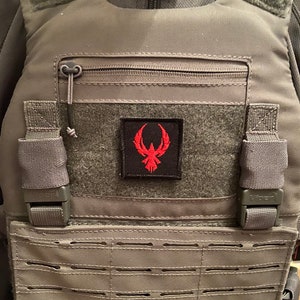 May include: A gray tactical vest with a zippered pocket and a red and black patch with a bird design. The vest has a molle system with multiple rows of webbing.