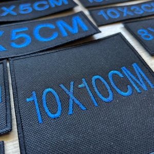 Peut inclure: Un ensemble de patchs en tissu noir avec du texte brodé bleu indiquant différentes tailles en centimètres. Les patchs comprennent les tailles 12x5cm, 10x3cm, 8x5cm et 10x10cm.