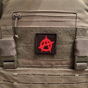 Puede incluir: Un chaleco táctico gris con un parche rojo con un símbolo de anarquía en la parte delantera. El chaleco tiene un bolsillo con cremallera y correas ajustables.