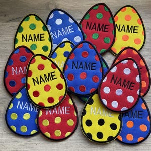 May include: A set of 10 colourful felt Easter egg-shaped patches with white polka dots and the word "NAME" in black lettering.