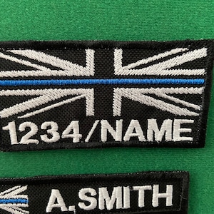Peut inclure: Un écusson brodé noir et blanc avec une ligne bleue au milieu. L'écusson présente un motif de drapeau britannique et le texte "1234/NAME".