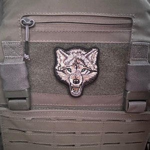 May include: A grey tactical vest with a wolf patch. The patch shows a detailed embroidered wolf head with a snarling expression. The vest has multiple pockets, a zip, and adjustable straps.