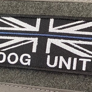 以下が含まれることがあります： 中央に青い線が引かれた黒と白の刺繍パッチ。パッチにはユニオンジャックの旗と「DOG UNIT」の文字が描かれています。