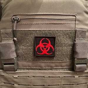 Puede incluir: Un chaleco táctico gris con un bolsillo con cremallera negro y un parche de símbolo de riesgo biológico rojo.