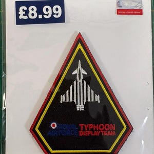 May include: A triangular embroidered patch featuring a white fighter jet silhouette on a black background with a yellow and red border. The text reads ROYAL AIR FORCE TYPHOON DISPLAY TEAM, accompanied by a small circular red, white, and blue roundel.