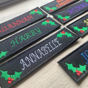 May include: A collection of rectangular black embroidered patches featuring names such as Susannah, Harry, Annabelle, and Anthony stitched in various colours, each decorated with green holly leaves and red berries at the corners.