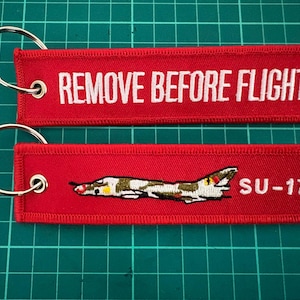 May include: Two rectangular red fabric keychains with silver metal rings. The top keychain features the white embroidered text REMOVE BEFORE FLIGHT. The bottom keychain displays an embroidered SU-17 aircraft graphic and the white text SU-17.