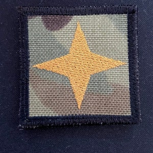 May include: A square patch with a camouflage background and a large, four-pointed, yellow star in the center.