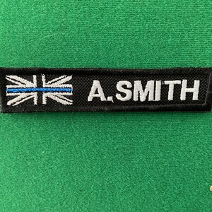 May include: A black and white embroidered patch with a blue line across the Union Jack flag and the text "A. SMITH" in white.