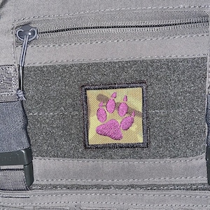 Peut inclure: Un sac à dos tactique gris avec un patch en forme de patte rose sur le devant. Le patch est cousu sur un panneau en velcro noir.