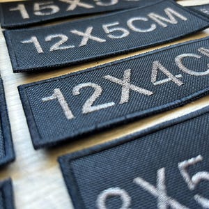 May include: Close-up of black fabric patches with white embroidered text. The patches show different sizes, including 12x5cm, 12x4cm, and 15x...