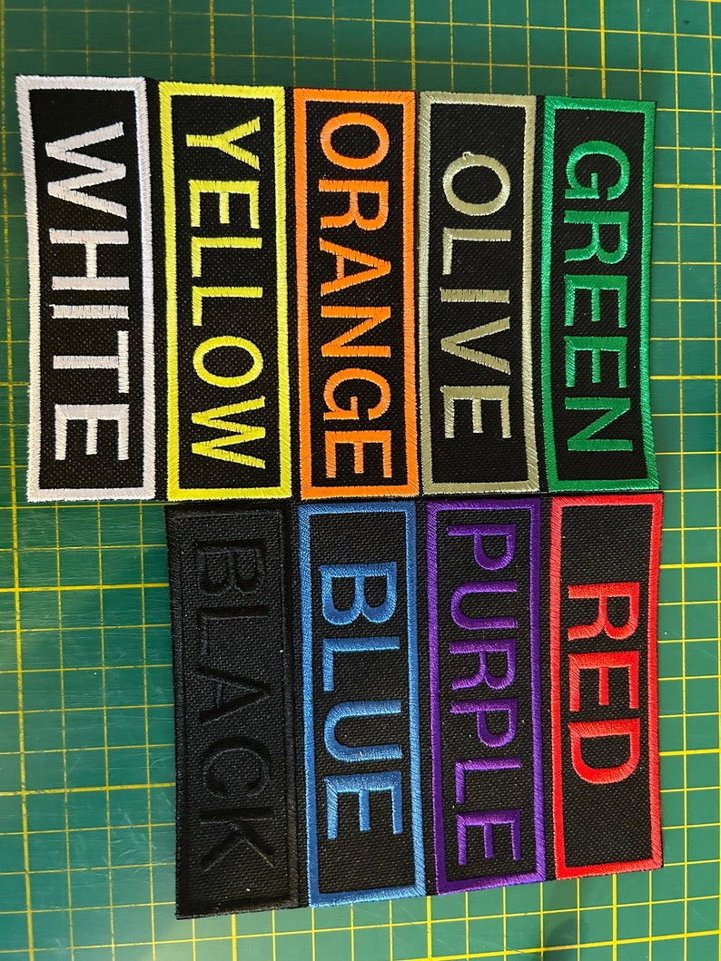 以下が含まれることがあります： さまざまな色の刺繍テキストが入った長方形のパッチのコレクション。パッチには、太字の大文字で「WHITE」、「YELLOW」、「ORANGE」、「OLIVE」、「GREEN」、「BLACK」、「BLUE」、「PURPLE」、「RED」という単語が表示されています。