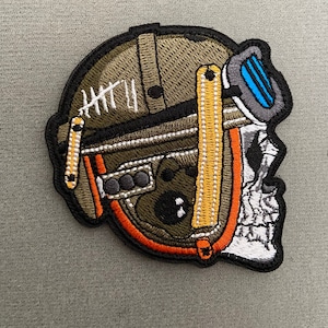 May include: An embroidered patch featuring a skull wearing a helmet with a visor. The helmet is green, orange and white with a yellow stripe on the strap and the text "HTTL" is visible.